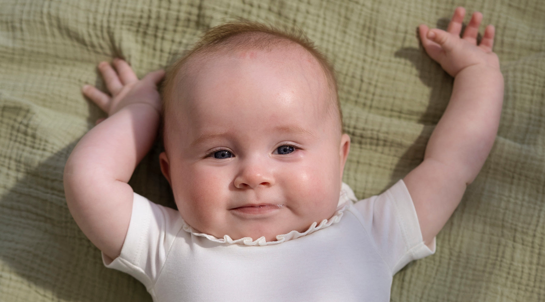Vrolijke baby in wit rompertje ligt op de rug op een groen deken met armpjes omhoog gestrekt