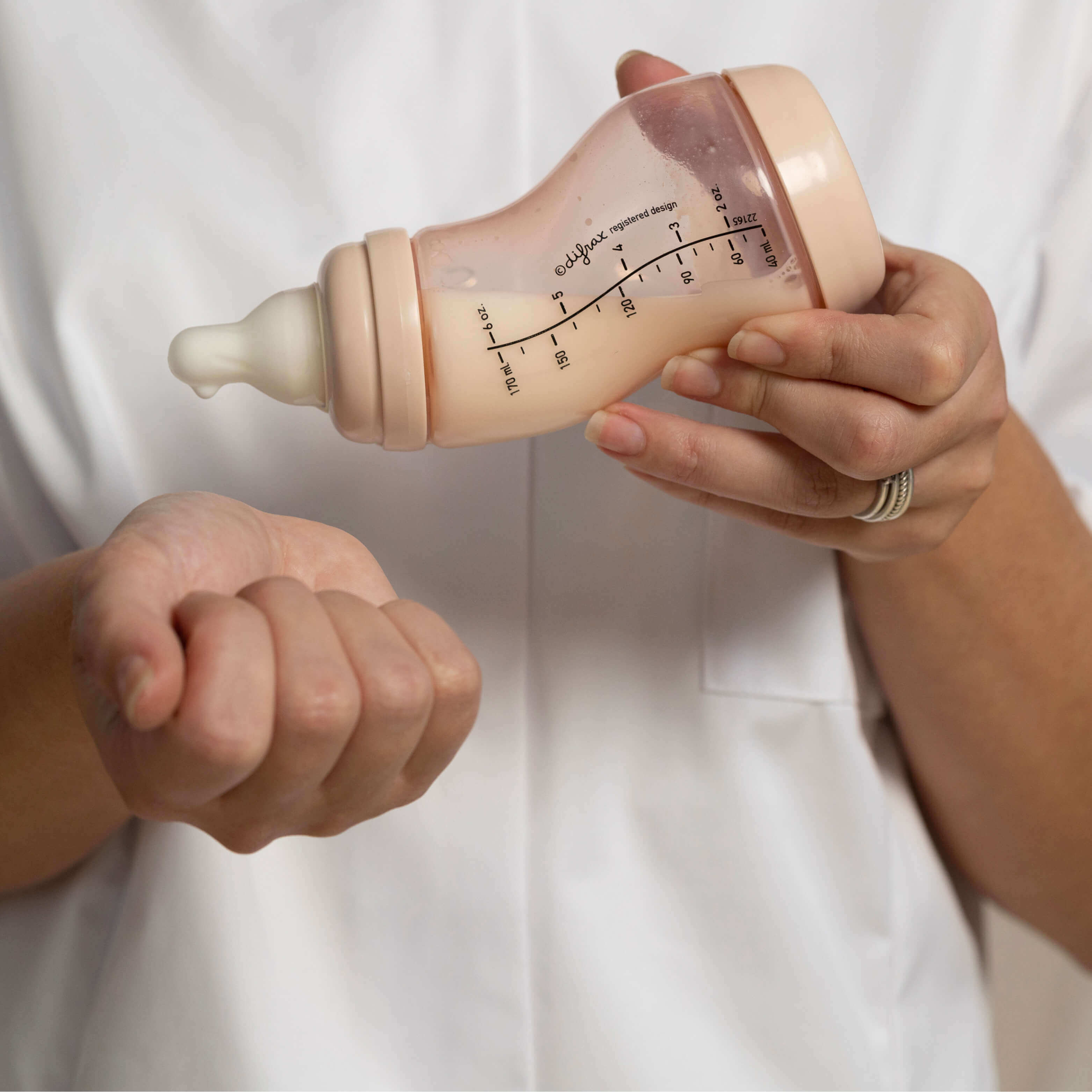 Close-up van handen die een Difrax S-fles in beige kleur vasthouden, met duidelijk zichtbare volumemaatverdeling en ergonomische grip