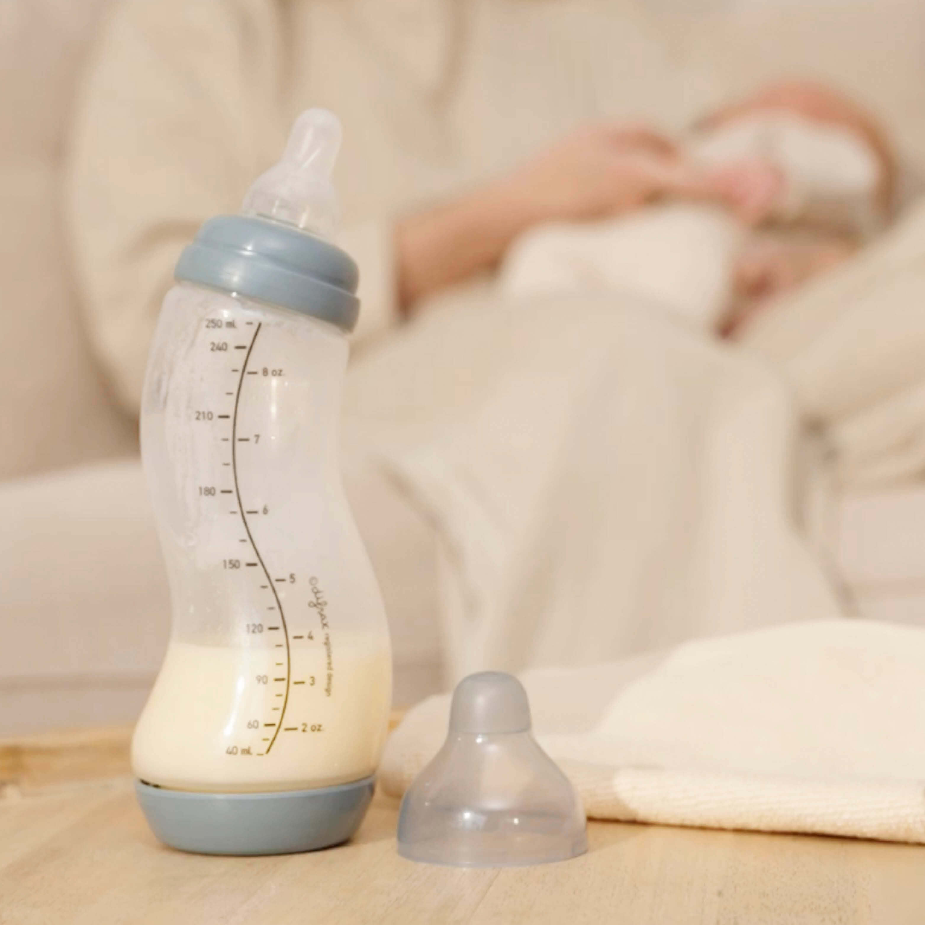 Stone grijze Difrax S-fles 250ml gevuld met moedermelk op houten ondergrond, slapende baby op zachte deken op achtergrond