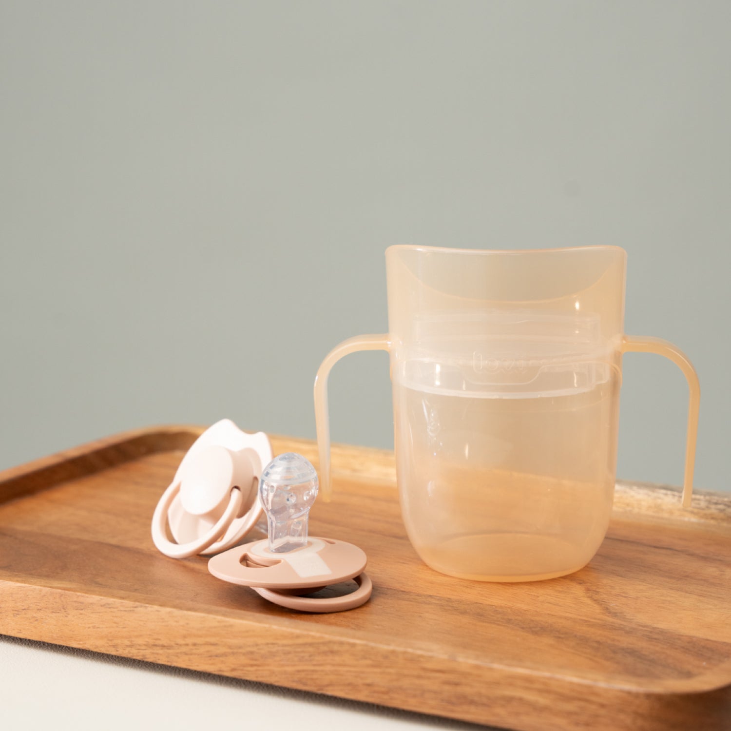 Difrax LOVI Genius oefenbeker in nude kleur op een tafel, naast twee dynamische fopspenen – stijlvol en functioneel babyproduct