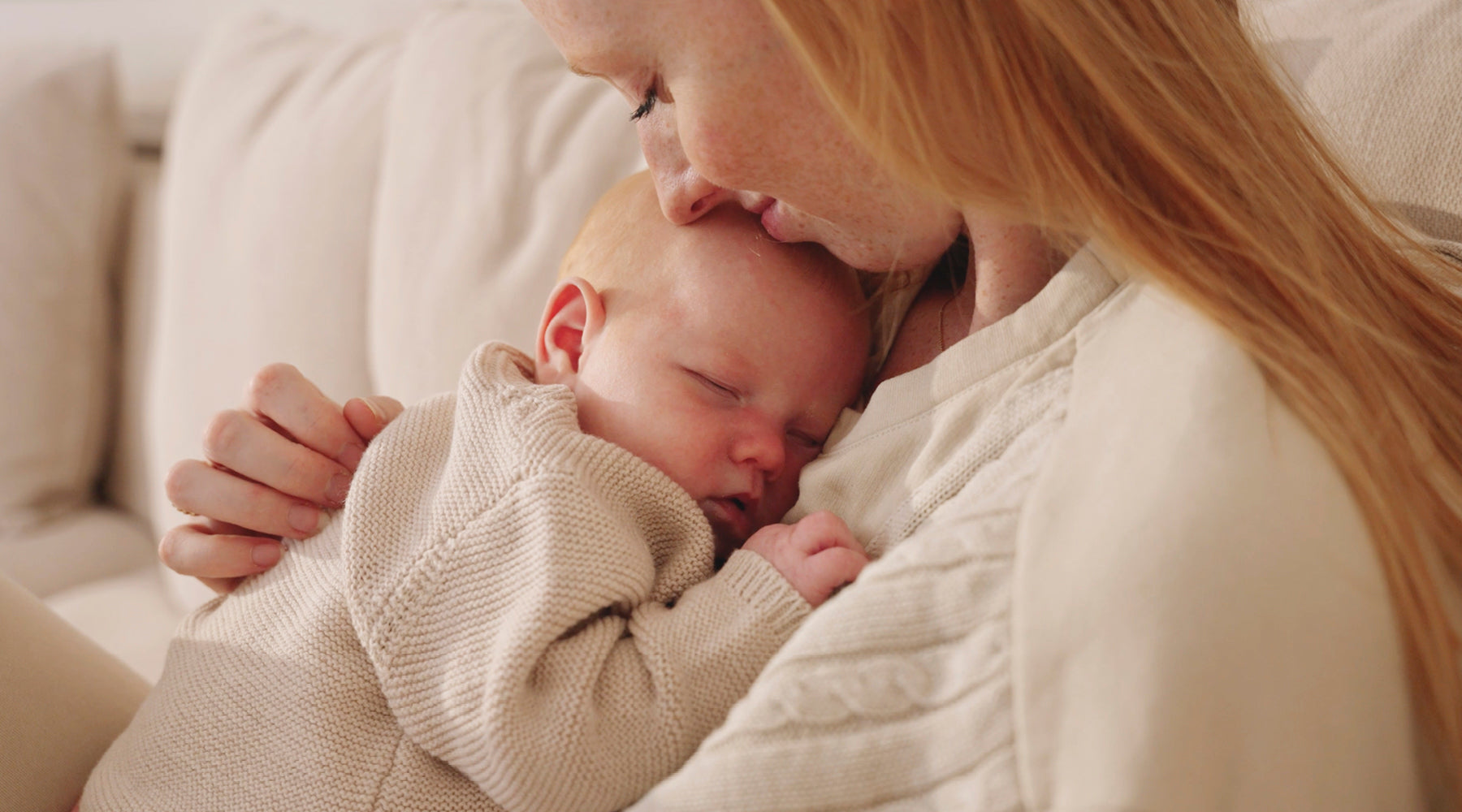 Moeder met rood haar houdt slapende baby in beige gebreid vestje liefdevol vast en kust het hoofdje