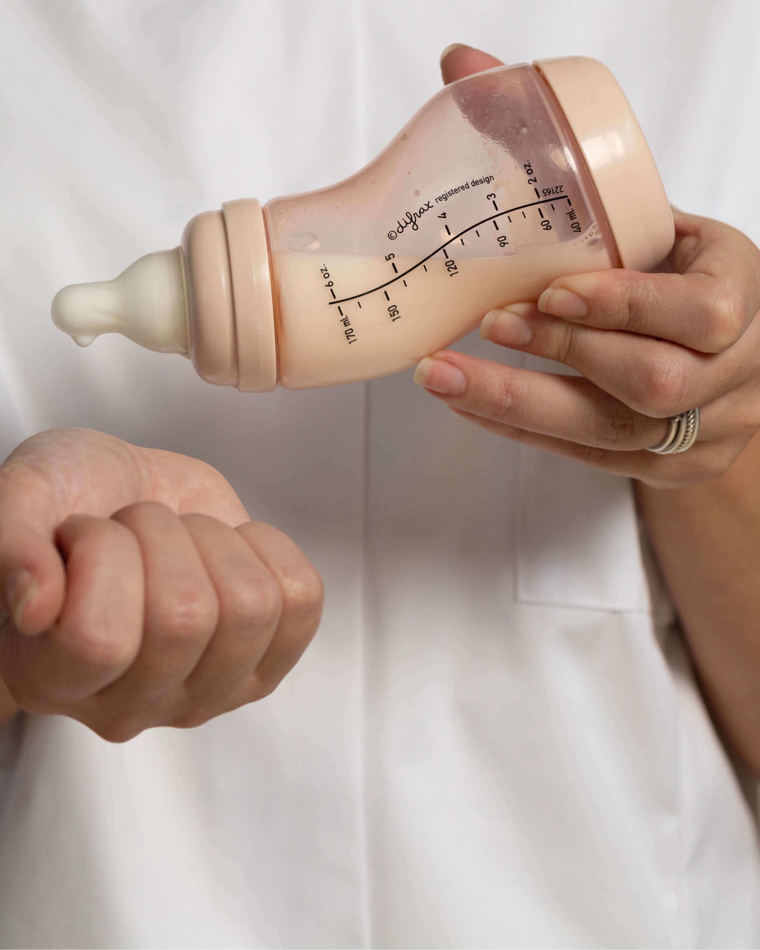 Close-up van handen die een Difrax S-fles in beige kleur vasthouden, met duidelijk zichtbare volumemaatverdeling en ergonomische grip