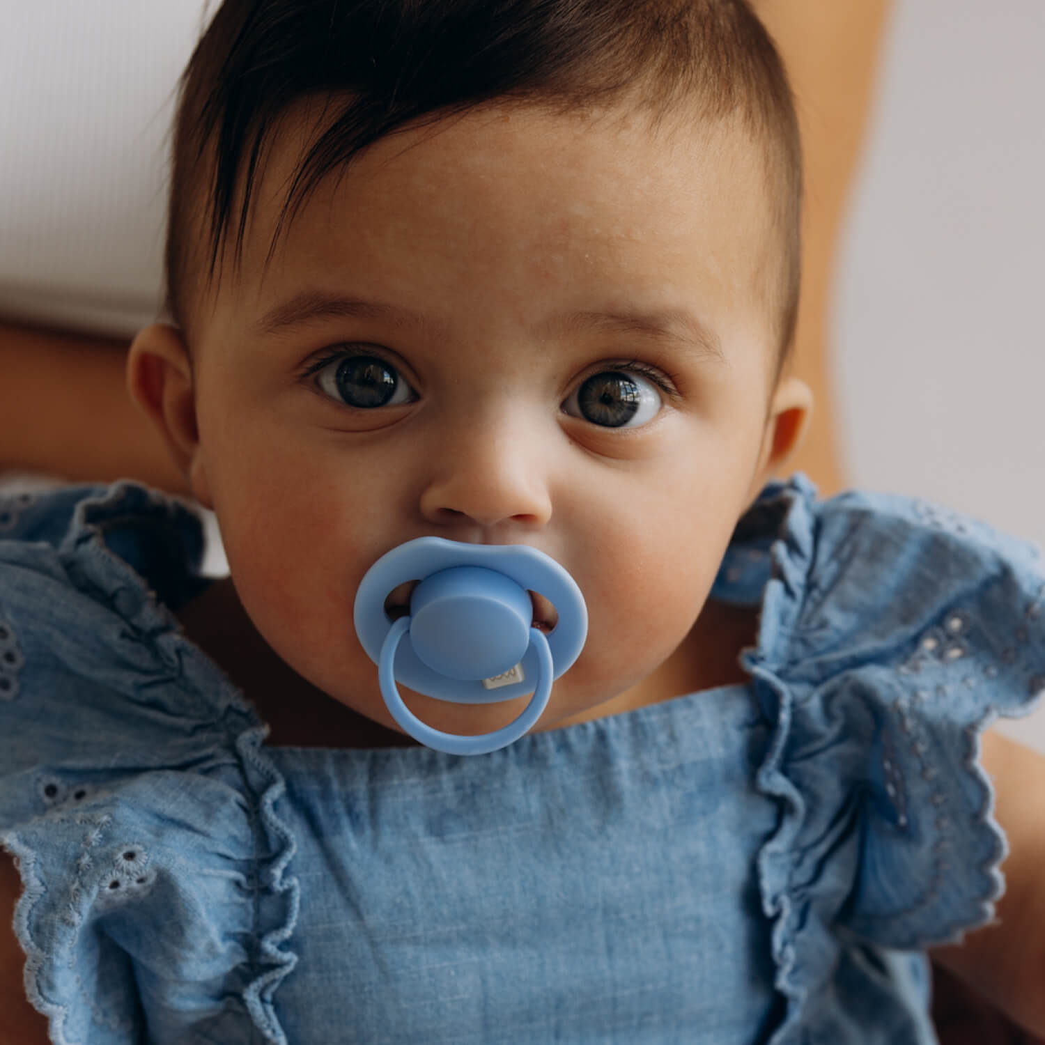 Donkerharige baby in een blauw jurkje met een lichtblauwe LOVI Dynamic fopspeen (Tutti Frutti, Ultramarine Blue)